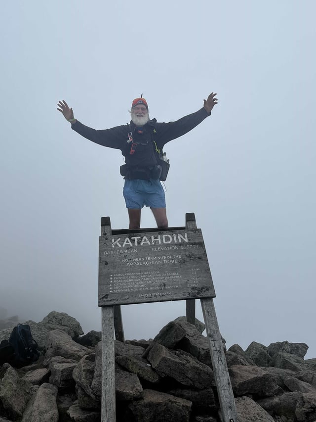 Jimmy, trail name Rocketman, with his arms raised behind the Katahdin summit sign on the Appalachian Trail